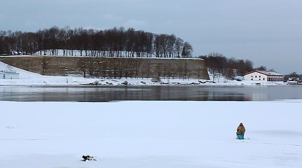 Mobility: The video Winter (2013) by Eva Engelbert was made in collaboration with sociologist Alena Pfoser. It  shows the paradoxes of a “twinned city” that is divided by a significant political border. It is not only a river that runs between Schengen and the Russian Federation, between Navra and Ivangorod, it also traces the hardship involved in overcoming an abstract entity in daily life. One inhabitant makes the point by telling of the difficulties she faces when visiting her husband on the other side of the river. On the other hand others cash in on it by smuggling goods past customs—even if it is only tea hidden in a pair of boots that are allegedly in need of immediate repair. Eva Engelbert & Alena Pfoser, still from 